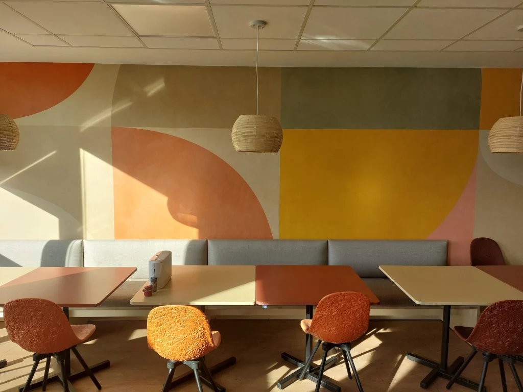 Cafétéria au style art déco avec un mur aux couleurs douces et géométriques ; au premier plan du mobilier écoresponsable.