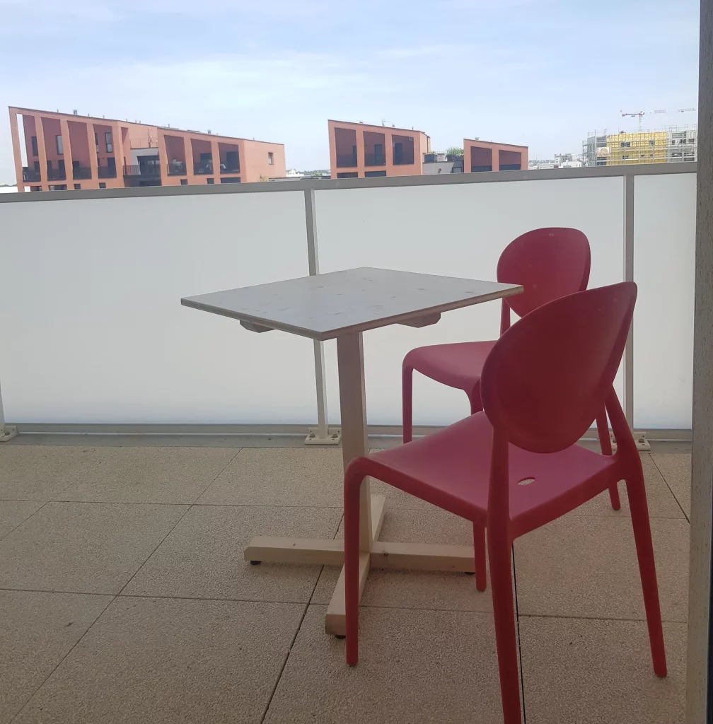 Deux chaises rouges et une table en bois de réemploi sont au premier plan de l'image, situés sur un balcon fermé agréable et paisible.
